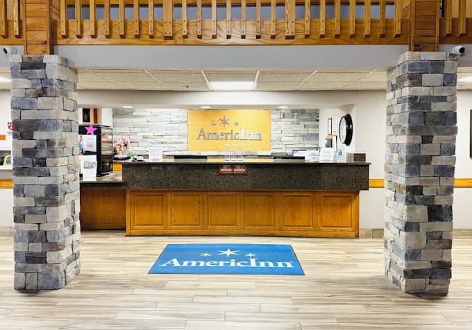 AmericInn lobby featuring a wooden reception desk, stacked stone pillars, and a branded floor mat.